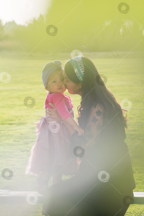 Скачать Молодая мать с дочерью на фоне зеленой лужайки. Девочка обнимается с маленькой девочкой и улыбается. Эффект боке и размытие переднего плана. фотосток Ozero