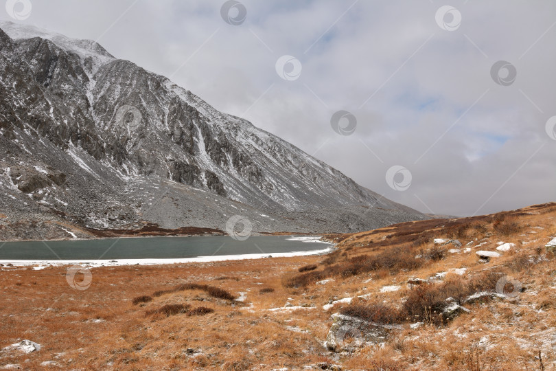 Скачать Фрагмент большого озера со льдом по берегам у подножия высокой скалы, припорошенный первым снегом в пасмурный осенний день. фотосток Ozero