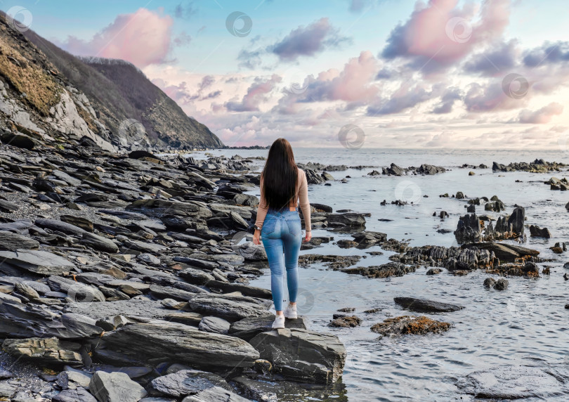Скачать Молодая стройная женщина прогуливается по пляжу с черными камнями и смотрит на закат фотосток Ozero