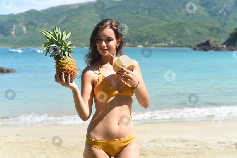 Скачать Молодая женщина в желтом купальнике пьет кокосовое молоко и держит в руках ананас на песчаном пляже фотосток Ozero