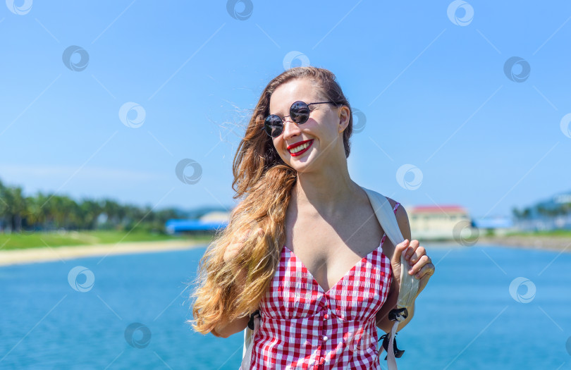 Скачать Яркая белокурая молодая женщина стоит с рюкзаком у моря летом фотосток Ozero