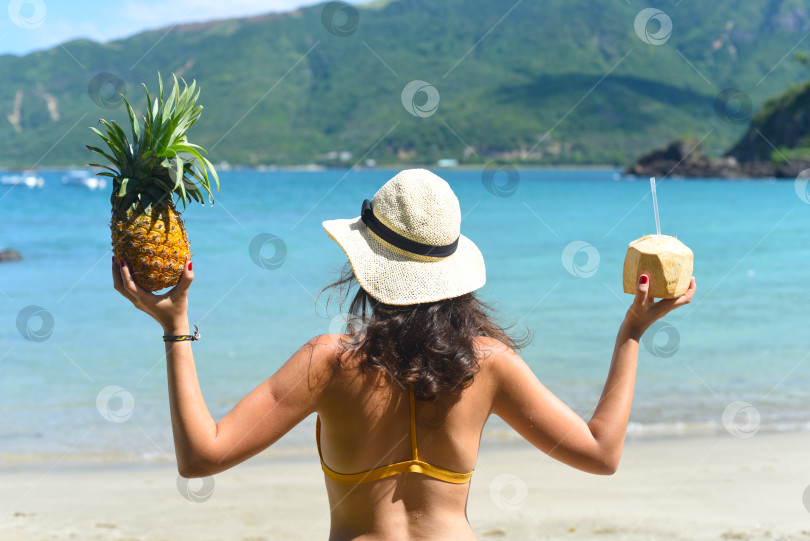 Скачать Молодая женщина в желтом купальнике держит кокос и ананас на песчаном пляже фотосток Ozero