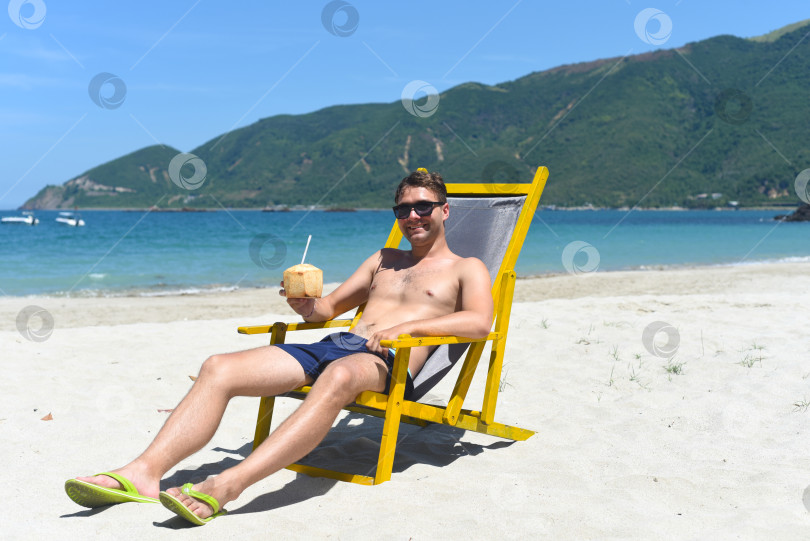 Скачать Молодой счастливый мужчина пьет кокосовый сок, сидя в шезлонге на пляже Южно-Китайского моря во Вьетнаме фотосток Ozero