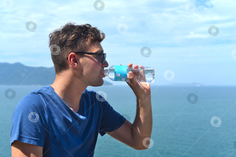 Скачать Молодой человек пьет воду из бутылки на фоне моря и голубого неба фотосток Ozero