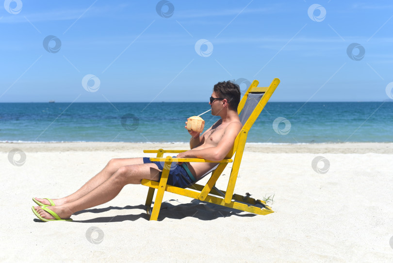 Скачать Молодой счастливый мужчина пьет кокосовый сок, сидя в шезлонге на пляже Южно-Китайского моря во Вьетнаме фотосток Ozero