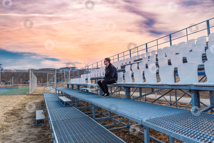 Скачать Один молодой человек сидит на пустой футбольной трибуне на улице в лучах заходящего солнца фотосток Ozero