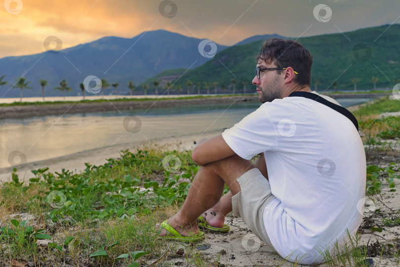 Скачать Расстроенный мужчина сидит на пляже и наблюдает за закатом в тропиках Вьетнама фотосток Ozero