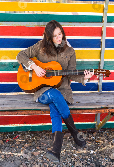 Скачать Молодая женщина играет на гитаре и смеется, сидя на скамейке на красочном фоне фотосток Ozero
