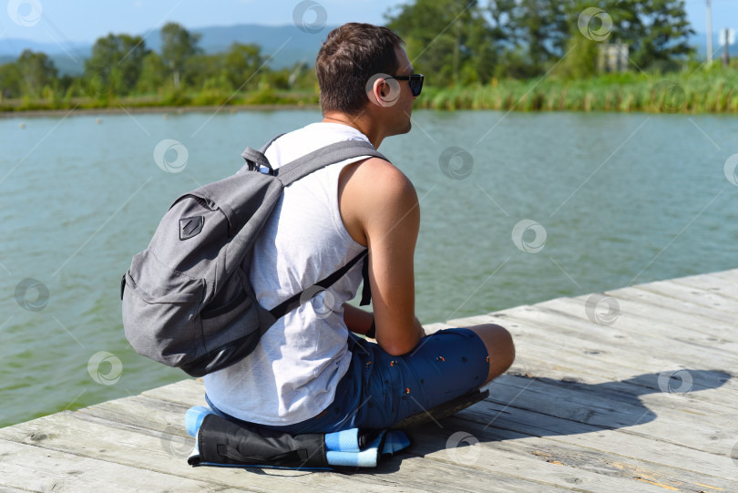 Скачать Молодой человек с рюкзаком отдыхает и сидит на пирсе пруда фотосток Ozero