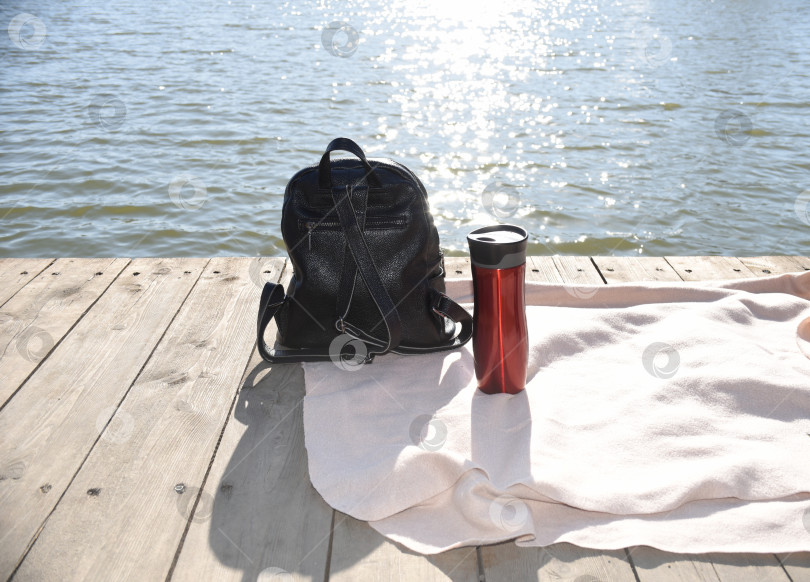 Скачать Черный рюкзак и красный термос с горячим чаем на пледе на деревянном пирсе на фоне японского моря и солнечных лучей. путешествия и пикники фотосток Ozero