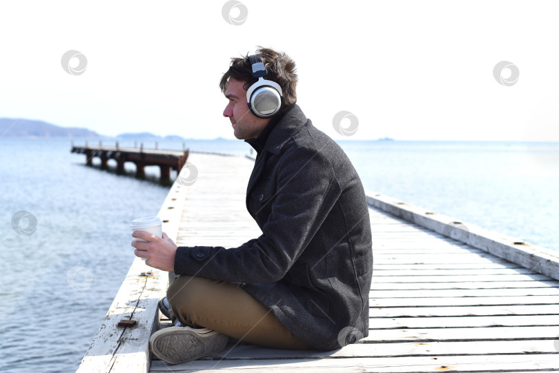 Скачать Молодой человек слушает музыку и пьет кофе, сидя в одиночестве на пирсе в солнечный день фотосток Ozero