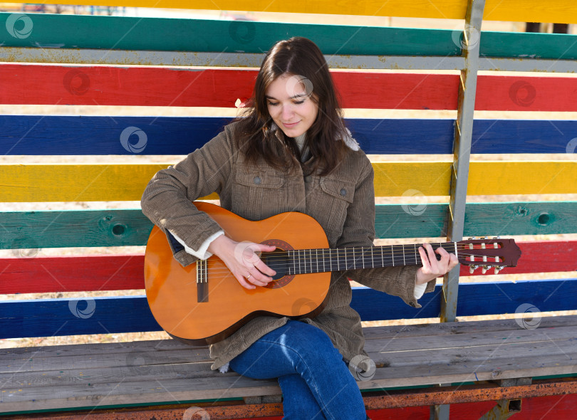 Скачать Молодая женщина играет на гитаре и смеется, сидя на скамейке на красочном фоне фотосток Ozero