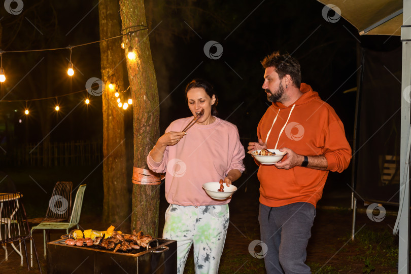 Скачать Супружеская пара, мужчина и женщина, едят сосиски-барбекю ночью возле гирлянд молний на заднем дворе фотосток Ozero