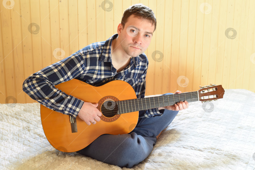 Скачать Молодой человек играет на гитаре, сидя на кровати в выходные утром. смотрит в камеру фотосток Ozero