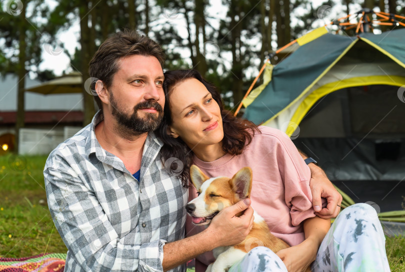 Скачать Молодая супружеская пара со своей собакой корги сидит в лесу возле палатки и смотрит на закат фотосток Ozero