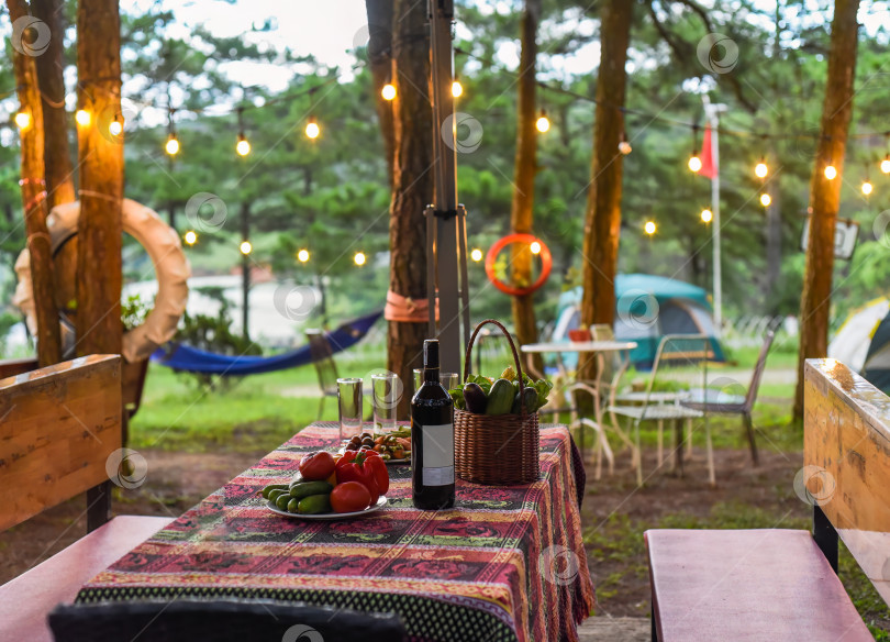 Скачать накрытый стол с бутылкой вина, овощами и бокалами рядом с гирляндами, гамаком и палаткой в лесу во время вечернего похода фотосток Ozero