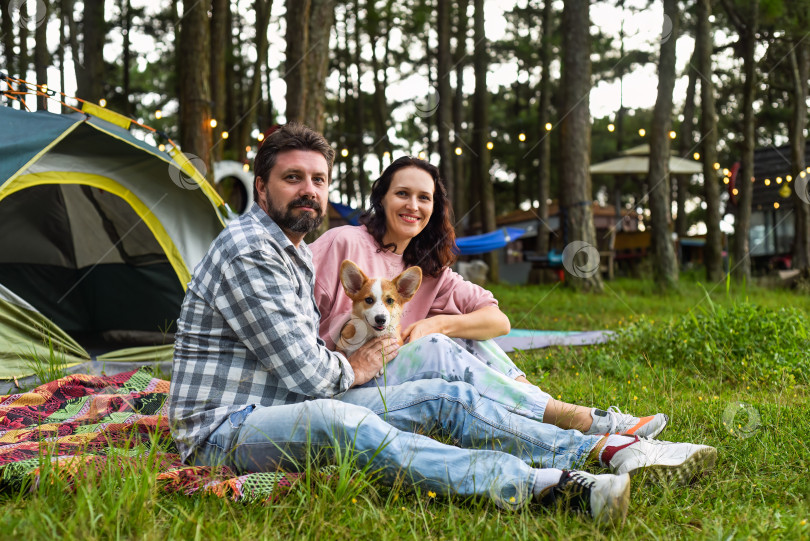 Скачать Молодая супружеская пара со своей собакой корги сидит в лесу возле палатки и смотрит на закат фотосток Ozero