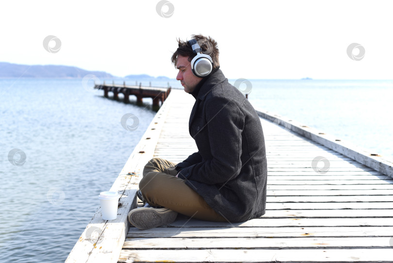 Скачать Молодой мрачный человек слушает музыку, сидя в полном одиночестве на пирсе в ветреный весенний день. фотосток Ozero