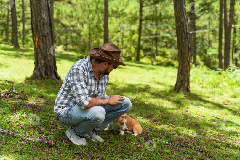 Скачать Молодой человек в шляпе трогает и играет со своей собакой корги в лесу фотосток Ozero