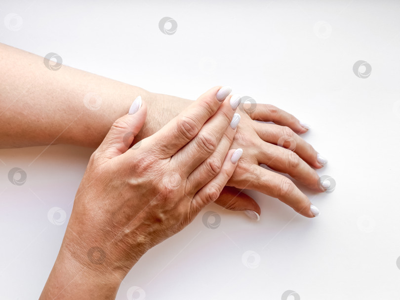 Скачать Руки женщины средних лет с белыми наманикюренными ногтями нежно касаются друг друга на белом фоне, вид сверху. Композиция в виде плоского слоя. Уход за кожей и руками или концепция материнской любви. фотосток Ozero