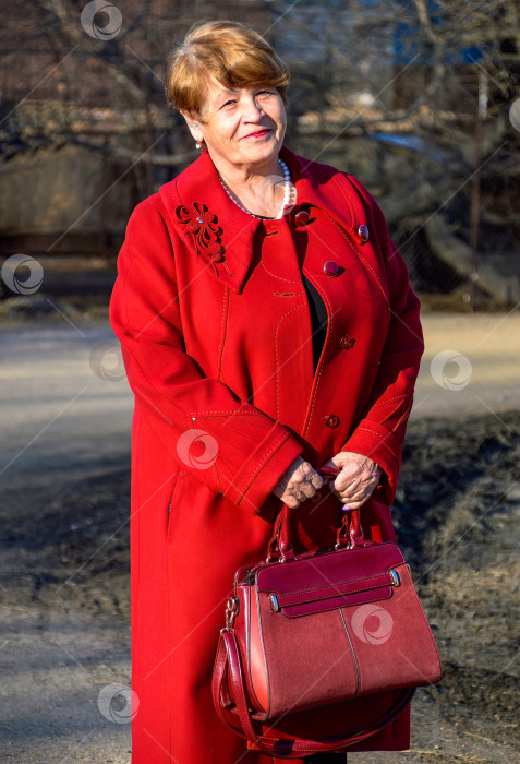 Скачать Портрет стильной 67-летней женщины в ярко-красном пальто с бордовой сумкой, любезно и искренне улыбающейся весенним днем фотосток Ozero