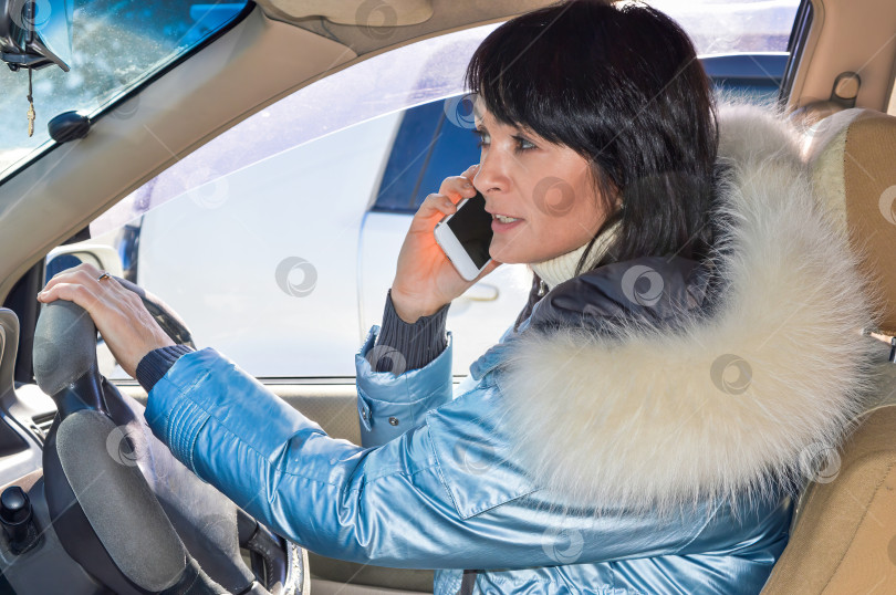 Скачать Молодая привлекательная женщина в куртке, смотрящая в сторону, ведет машину и разговаривает по телефону зимним днем фотосток Ozero