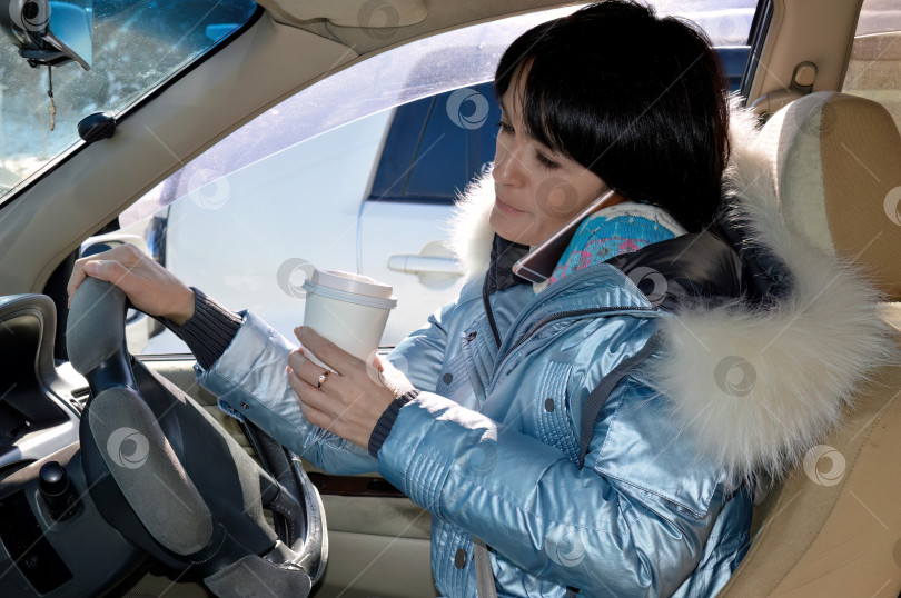 Скачать Молодая привлекательная женщина в куртке ведет машину, пьет кофе и разговаривает по телефону зимним днем фотосток Ozero