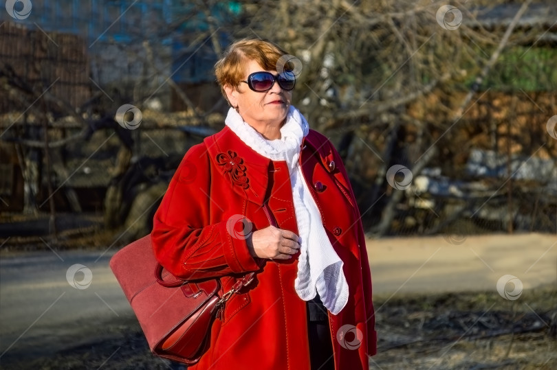 Скачать Портрет стильной 67-летней женщины в ярко-красном пальто, с бордовой сумкой, солнцезащитными очками и белым шарфом в весенний день фотосток Ozero