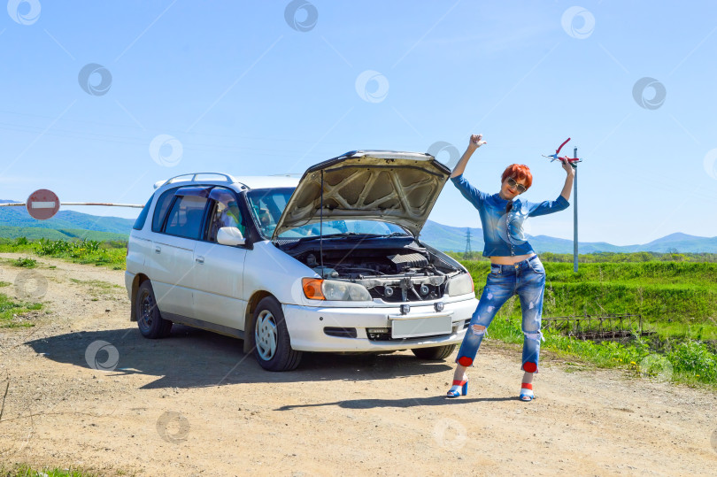 Скачать Рыжеволосая женщина с отверткой и плоскогубцами на высоких каблуках стоит возле разбитой машины с открытым капотом на проселочной дороге и шлагбаумом со знаком "стоп". фотосток Ozero