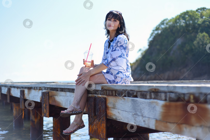 Скачать 45-летняя русская женщина пьет лимонад и сидит на пирсе фотосток Ozero