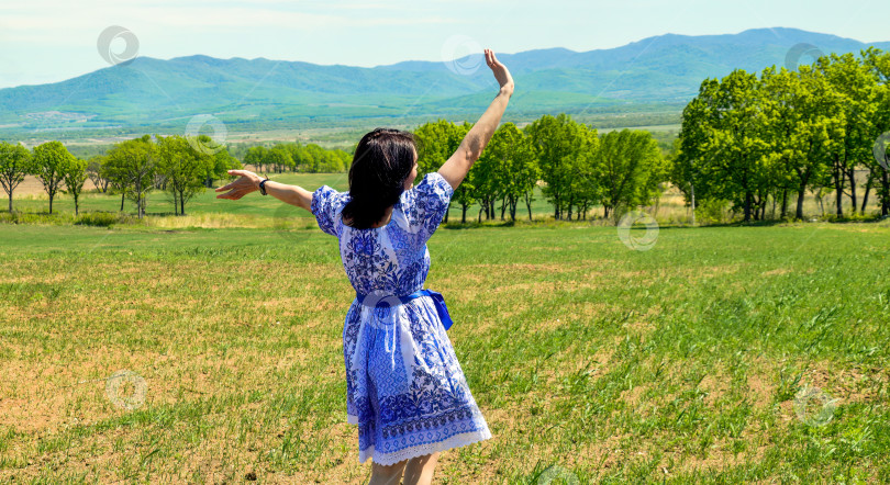 Скачать Русская молодая женщина в платье в славянском стиле бежит и машет рукой природе на фоне зеленого поля с деревьями. летнее время, концепция свободы фотосток Ozero