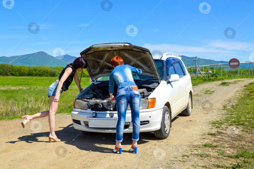 Скачать Рыжеволосая женщина с отверткой, на высоких каблуках, стоящая возле разбитой машины с открытым капотом, и брюнетка на проселочной дороге у шлагбаума со знаком "стоп". фотосток Ozero