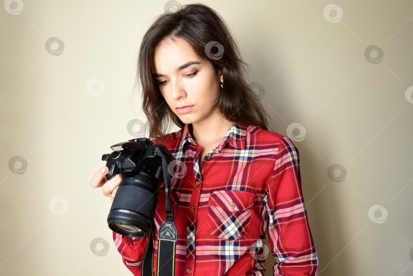 Скачать Уставшая серьезная женщина-фотограф в красной рубашке держит в руках фотоаппарат и рассматривает неудачные фотографии на бежевом фоне. Расстроена результатом. Место для копирования фотосток Ozero