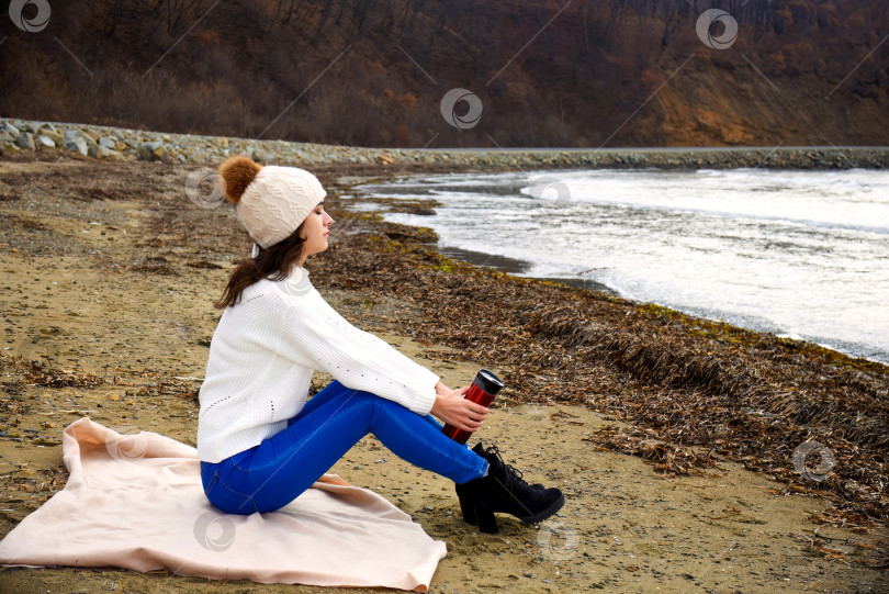 Скачать Молодая женщина в белом свитере, шапке с помпоном, сидит на берегу Японского моря с закрытыми глазами, вдыхая аромат зимнего воздуха. Она держит в руках термос с чаем. Скопировать место фотосток Ozero