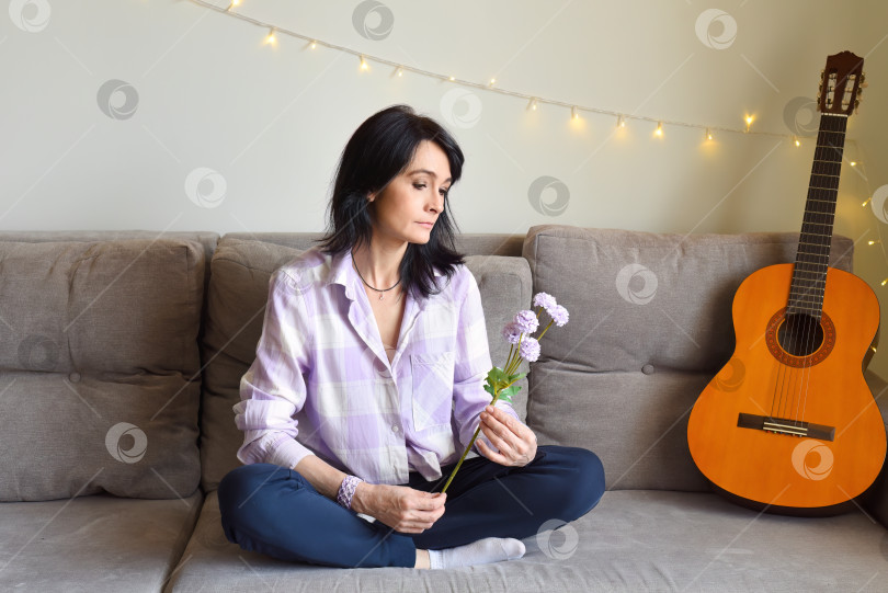 Скачать Расстроенная 45-летняя русская женщина смотрит на цветы сирени, сидя дома на диване. фотосток Ozero