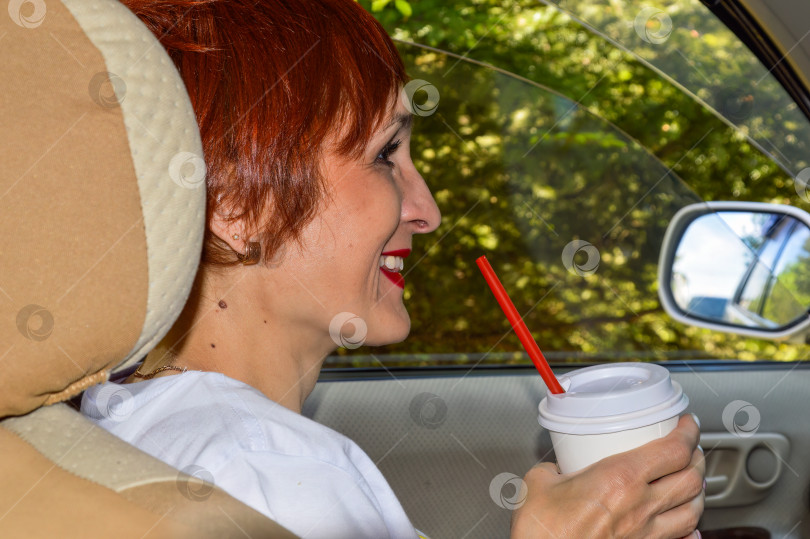 Скачать Веселая молодая женщина-пассажир с рыжими волосами сидит в машине, держа пластиковый стаканчик с кофе и улыбаясь во весь рот. Летние приключения фотосток Ozero
