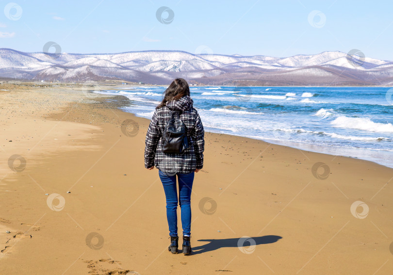 Скачать Молодая брюнетка с рюкзаком прогуливается по пляжу рядом со снежными горами и Японским морем. Одинокий путешественник. фотосток Ozero