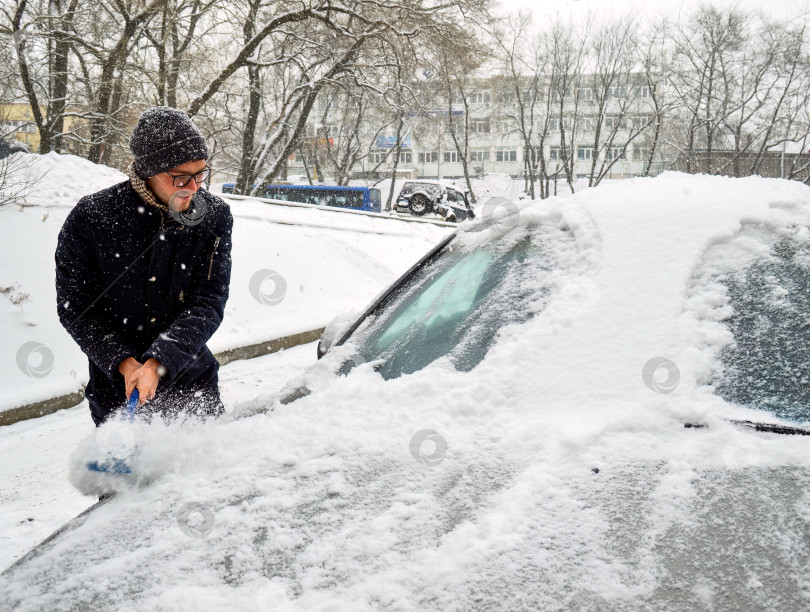 Скачать Молодой человек сметает снег с машины в снежный зимний день фотосток Ozero