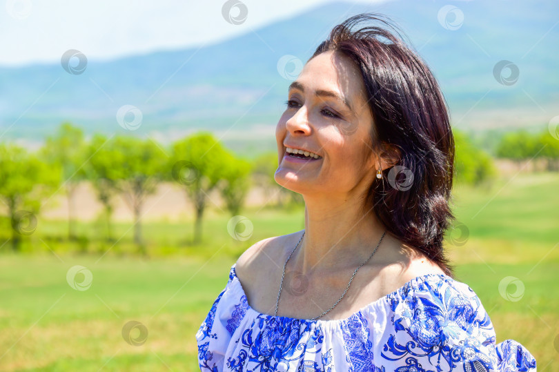 Скачать Портрет 45-летней белой брюнетки, смотрящей в камеру на зеленом поле на фоне гор и деревьев. Место для копирования фотосток Ozero