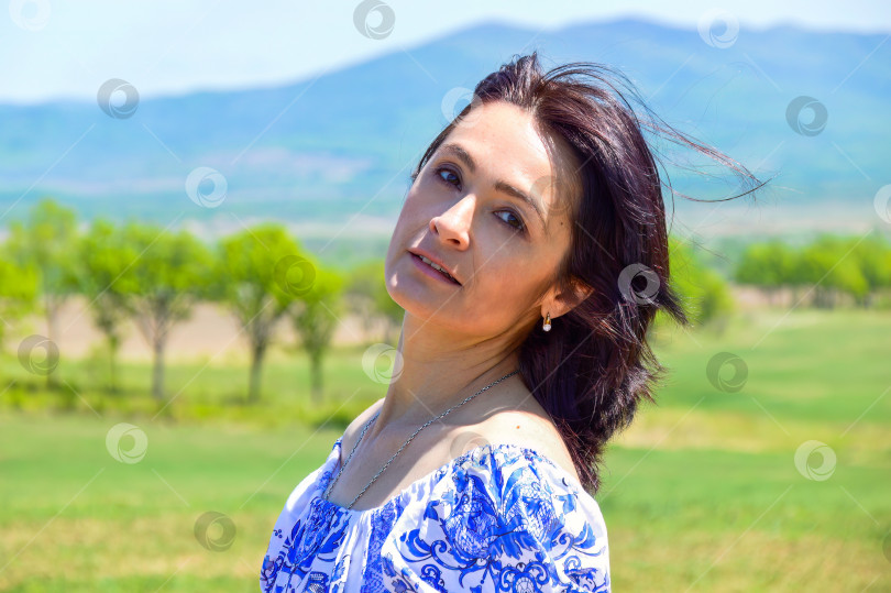 Скачать Портрет 45-летней белой брюнетки, смотрящей в камеру на зеленом поле на фоне гор и деревьев. Место для копирования фотосток Ozero