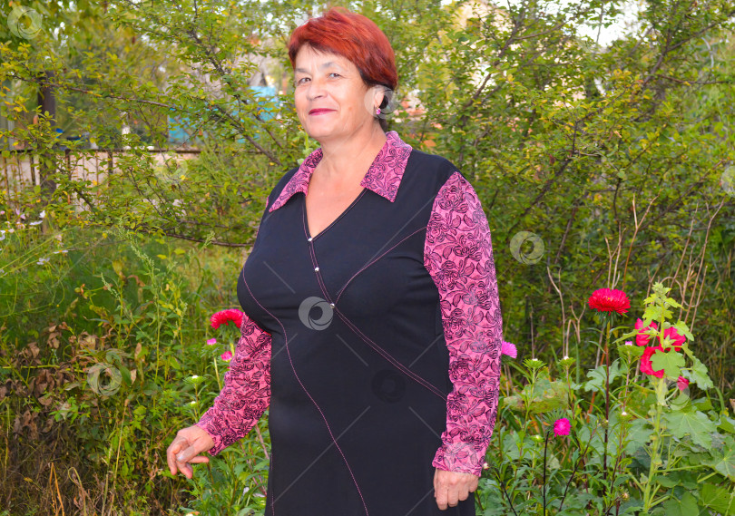 Скачать Пожилая симпатичная женщина в платье улыбается и стоит возле красивых розовых и красных астр. фотосток Ozero