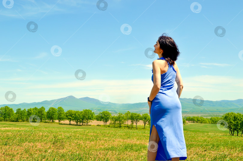Скачать Стройная 45-летняя улыбающаяся брюнетка в длинном синем платье с открытыми плечами на зеленом поле на фоне гор. Copy space фотосток Ozero