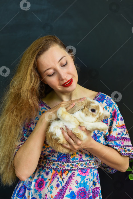 Скачать Красивая русская женщина с длинными светлыми волосами держит в руках домашнего кролика на фоне черной стены фотосток Ozero