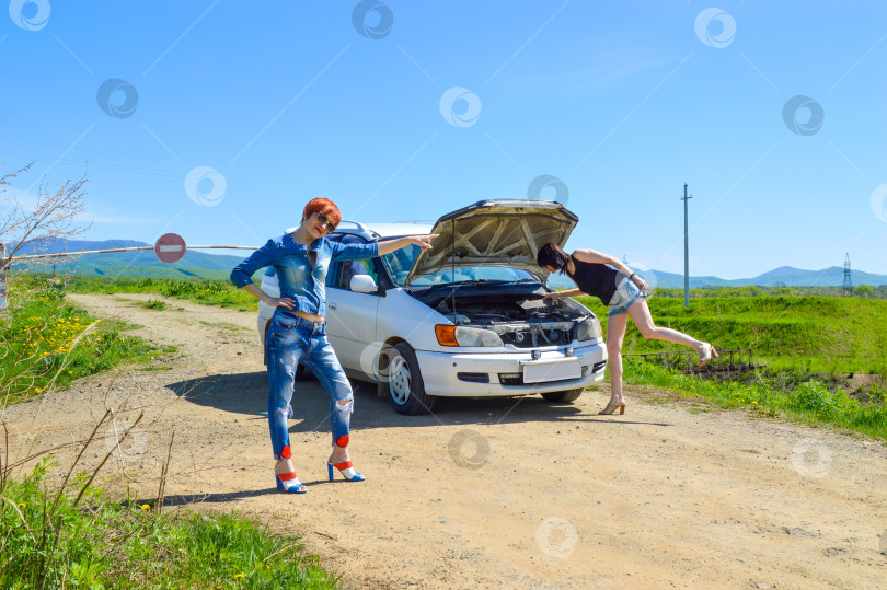 Скачать Рыжеволосая женщина, указывающая пальцем на разбитую машину с открытым капотом, и брюнетка на проселочной дороге и шлагбауме со знаком "стоп". фотосток Ozero