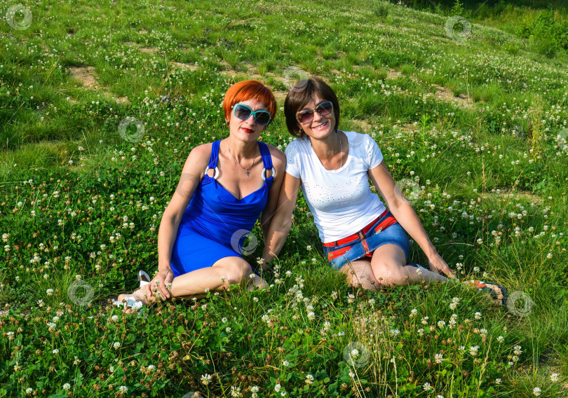 Скачать Две молодые красивые сестры в солнцезащитных очках сидят на траве и искренне улыбаются летним днем в России фотосток Ozero