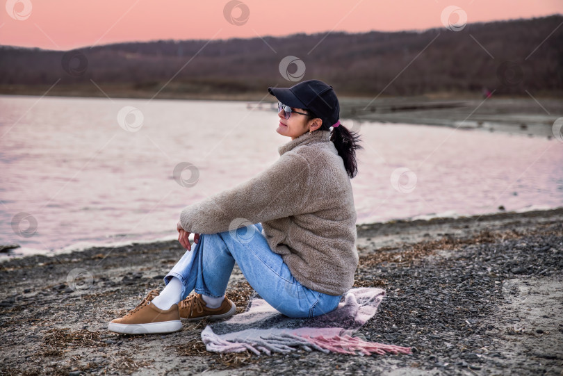Скачать 49-летняя русская женщина в куртке сидит на пляже и размышляет на фоне красного заката фотосток Ozero