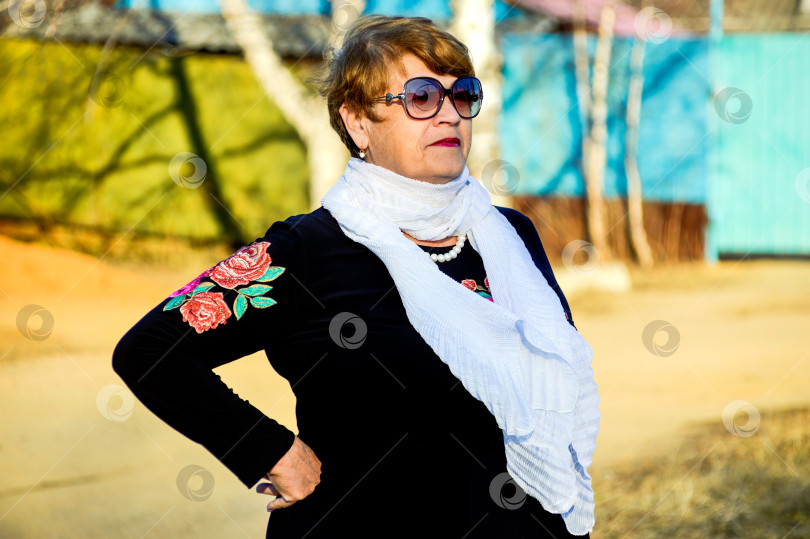 Скачать Портрет стильной 67-летней женщины в черной блузке с цветами и солнцезащитных очках, любезно и искренне улыбающейся весенним днем фотосток Ozero