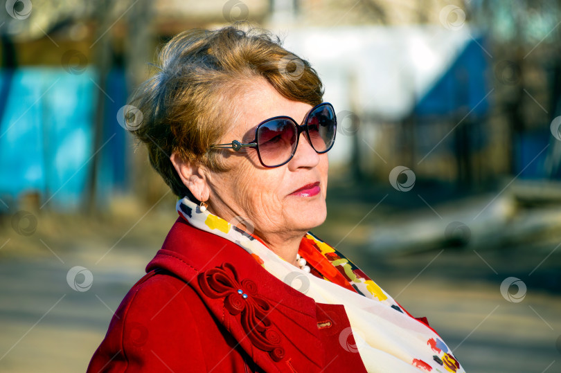Скачать Портрет стильной 67-летней женщины в ярко-красном пальто, солнечных очках и белом шарфе в весенний день фотосток Ozero