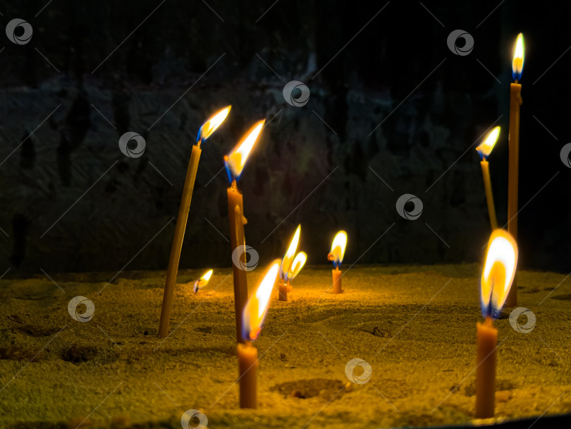 Скачать Зажженные свечи в подсвечнике, наполненном песком, в церкви на фоне темной стены. Религиозная или духовная церемония с мерцающим пламенем и теплым светом свечей. Зажженные свечи в церкви. фотосток Ozero