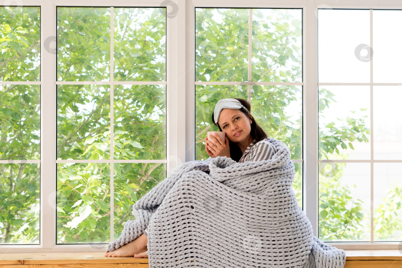 Скачать Расслабленная женщина сидит у окна с кружкой и одеялом. фотосток Ozero
